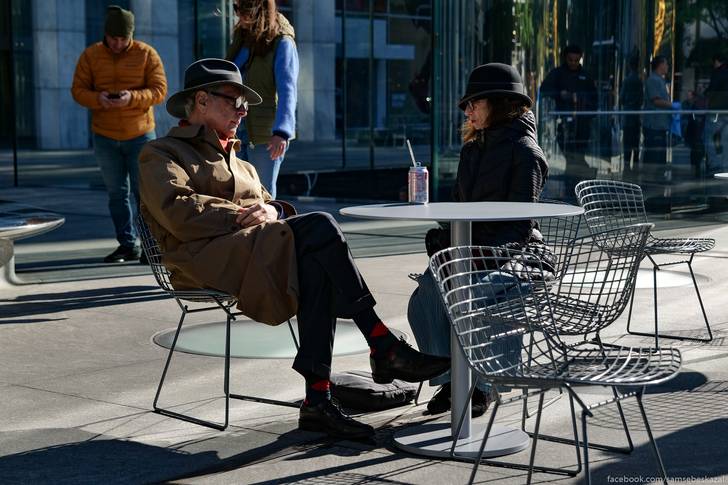 a man in a trench coat and a woman in a dark hat sit at a table in a pedestrian plaza